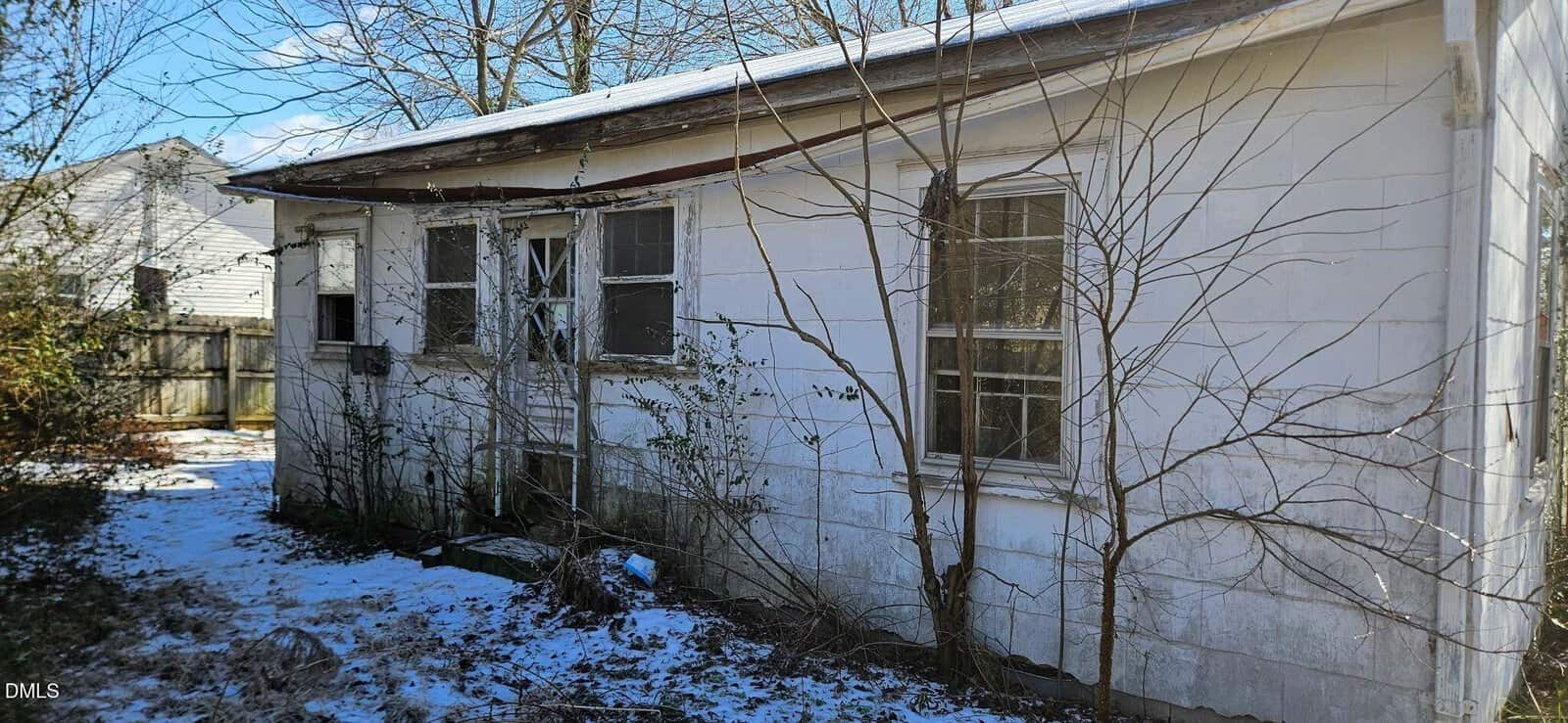 A small, weathered white house with peeling paint, broken windows, and overgrown leafless branches. Patches of snow cover the ground, and sunlight filters through the bare trees in the background.