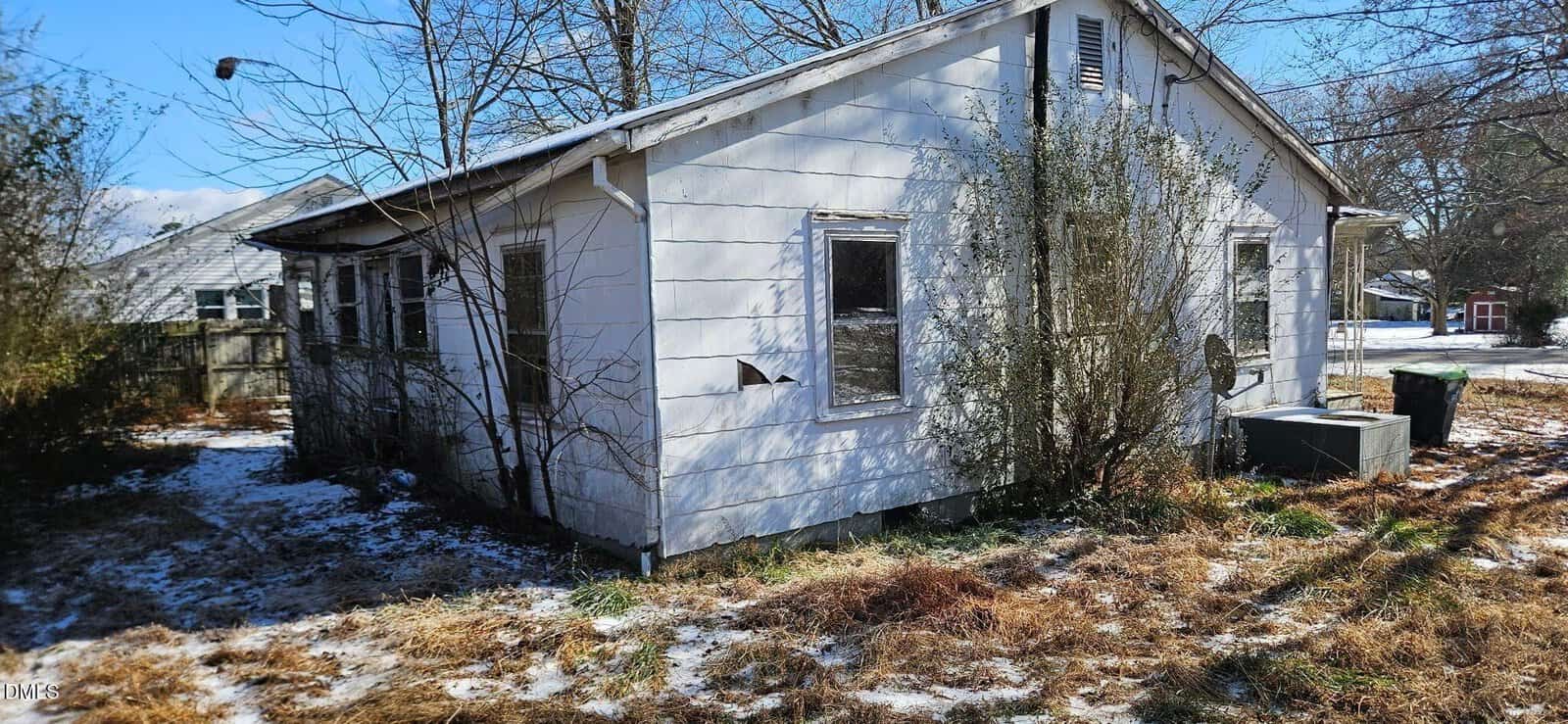 A small, weathered white house with peeling paint and overgrown bushes, sits on a patchy, snow-dusted yard under a clear blue sky. An air conditioning unit and trash bins are visible beside the house.