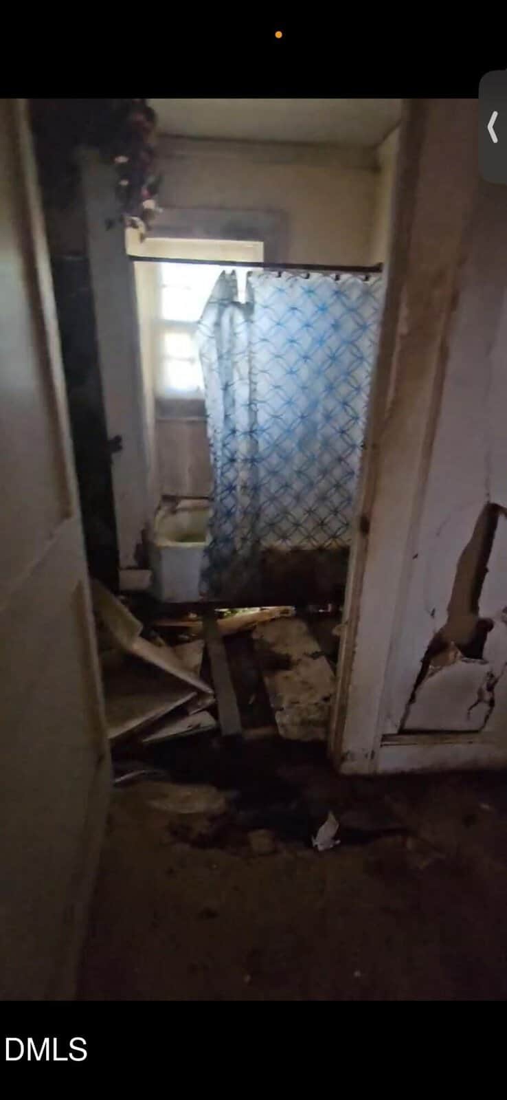A dimly lit, rundown bathroom with debris covering the floor, a toilet near a small window, and a blue-patterned shower curtain. The walls and door are damaged, showing holes and cracks.