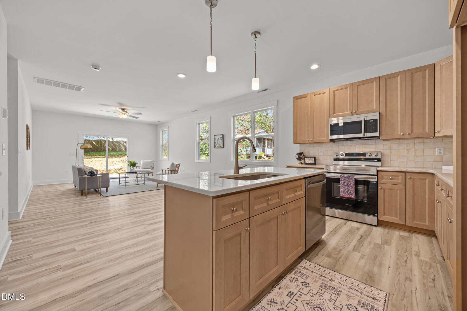 Modern open-concept kitchen and living room with light wood cabinets, stainless steel appliances, island with pendant lights, and large windows providing natural light. Light wood flooring throughout the space.