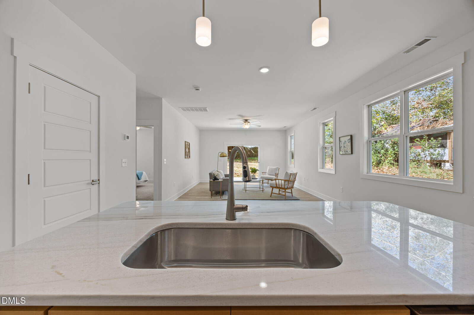 Modern open-concept living space with a kitchen island and sink in the foreground, pendant lights overhead, and a bright living area with large windows and contemporary furniture in the background.