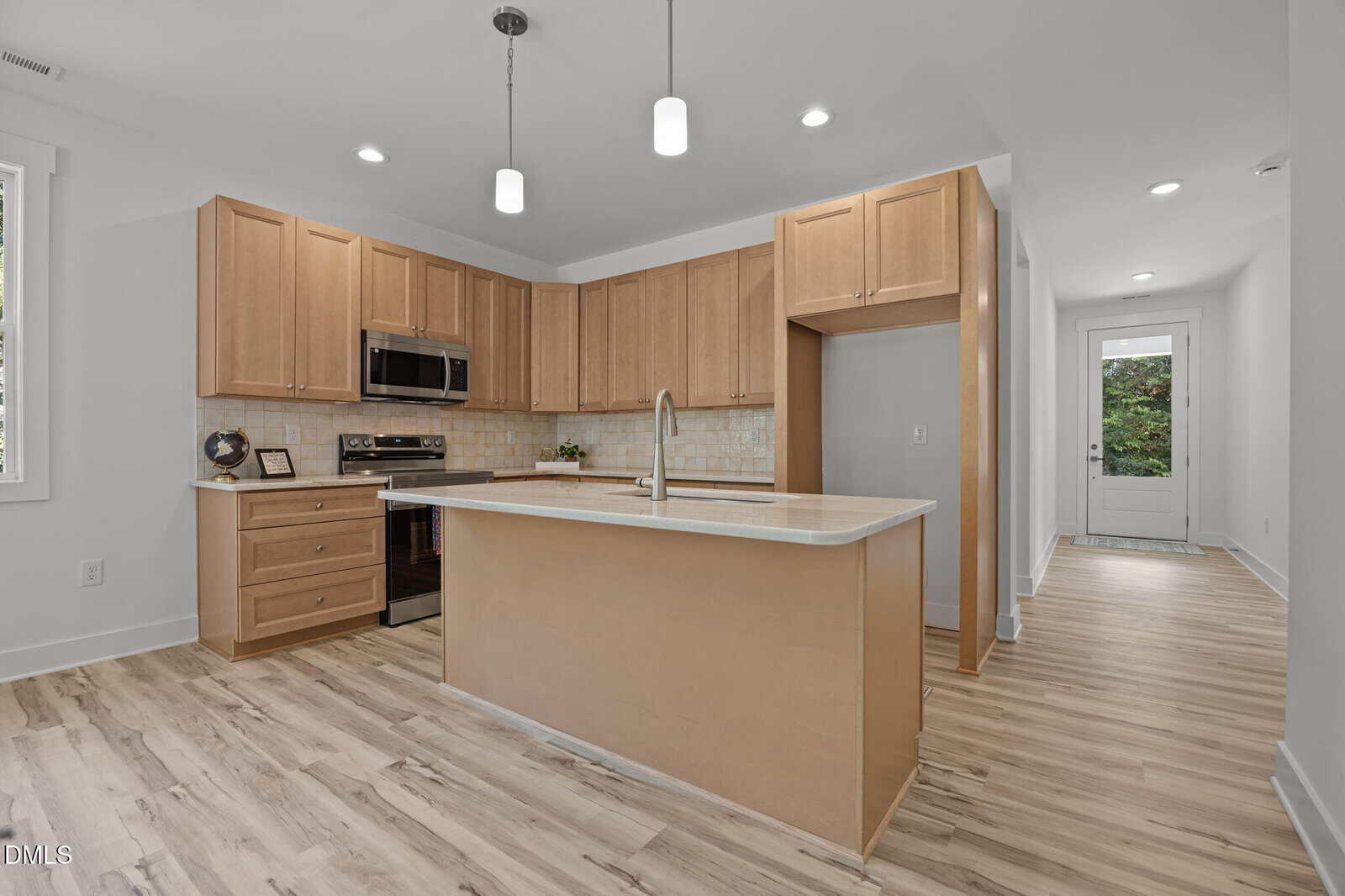 Modern kitchen with light wood cabinets, stainless steel appliances, a center island with a sink, light wood flooring, white walls, recessed ceiling lights, and a glass door leading outside.