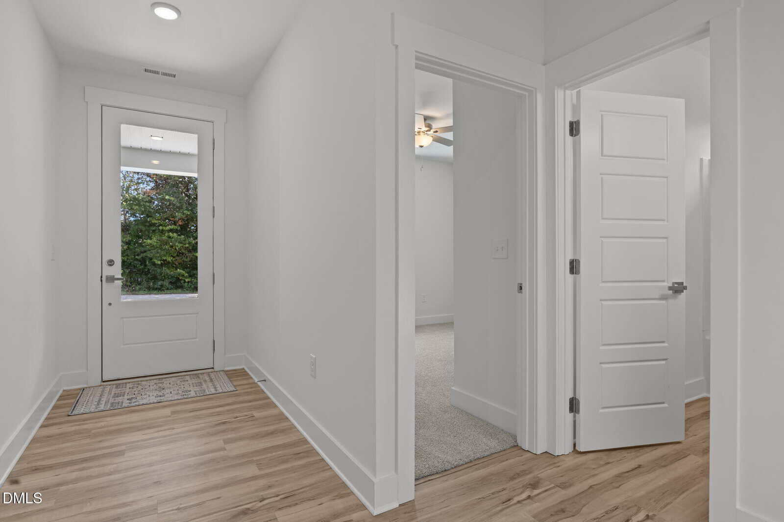 A bright, modern hallway with light wood flooring, white walls, and a glass door leading outside. Two open doorways on the right reveal carpeted rooms; one door is partially open. A rug lies inside the entrance.