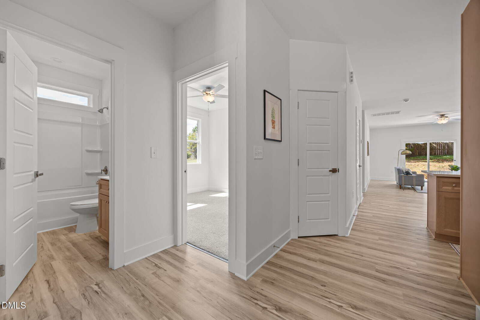 A bright hallway with light wood flooring leads to a bathroom, a carpeted bedroom, and a living area with large windows. White walls and doors create a clean, modern look.