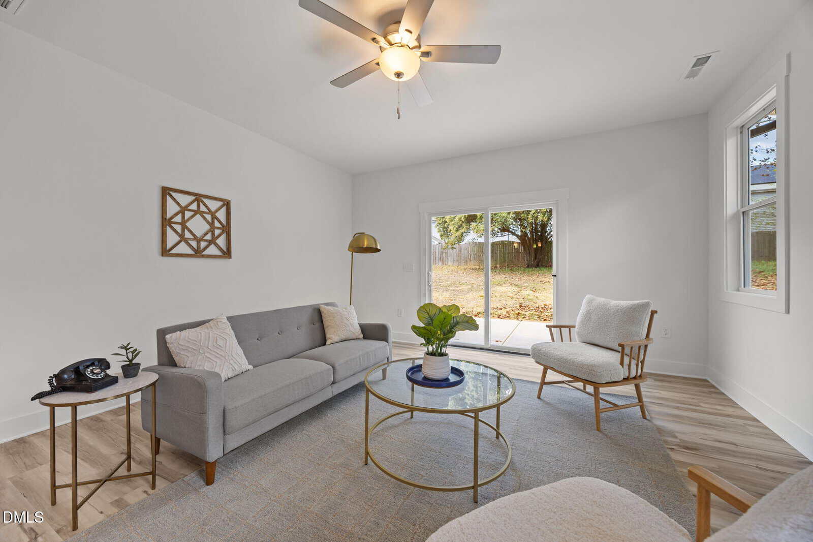 A bright living room with a gray sofa, light armchair, round coffee table, potted plant, floor lamp, wall art, and large windows with a sliding glass door overlooking a grassy backyard.