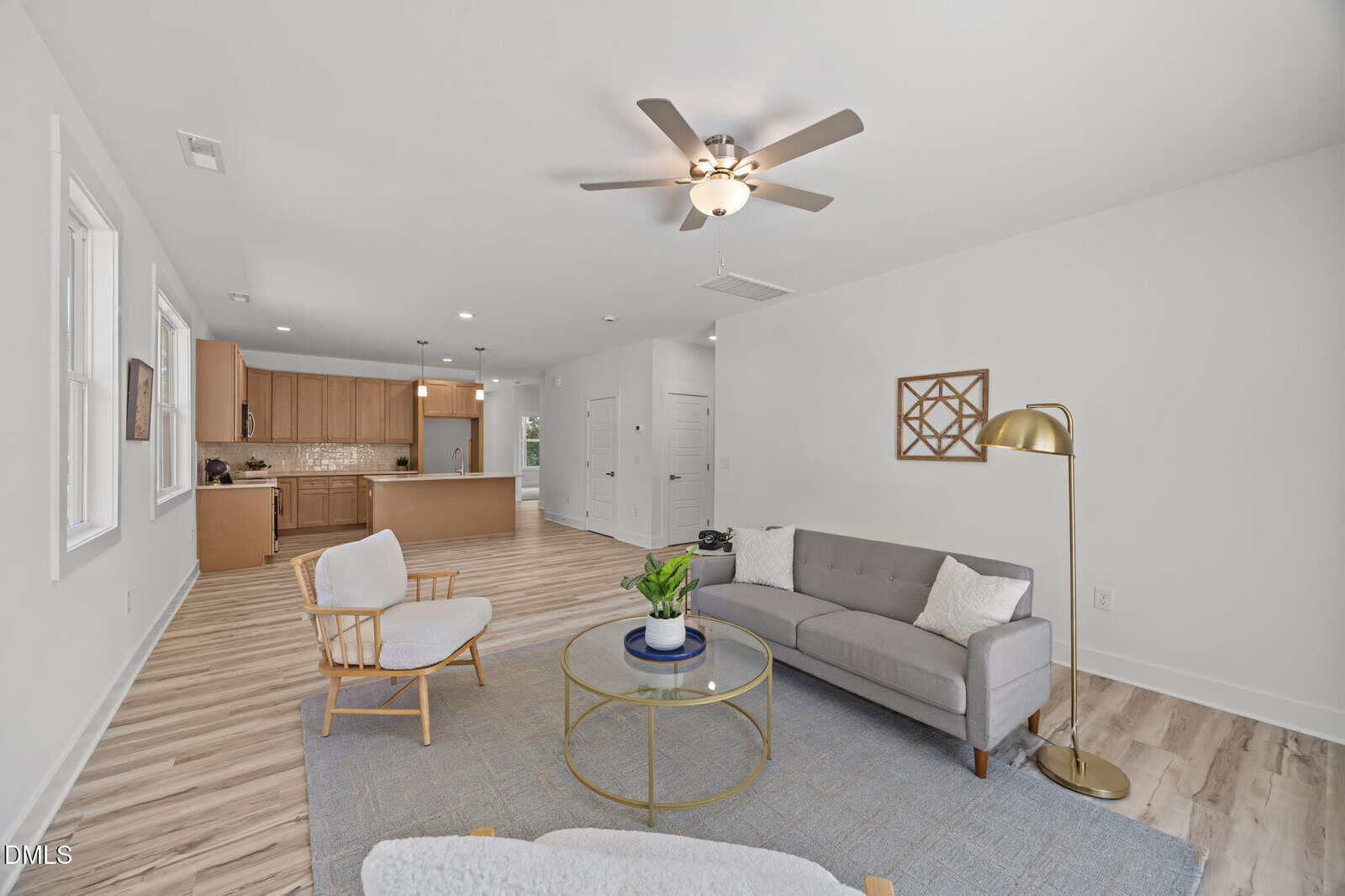 Open-concept living room and kitchen with light wood floors, gray sofa, white armchair, round glass coffee table, floor lamp, and modern light wood kitchen cabinets in the background. Neutral, airy decor.