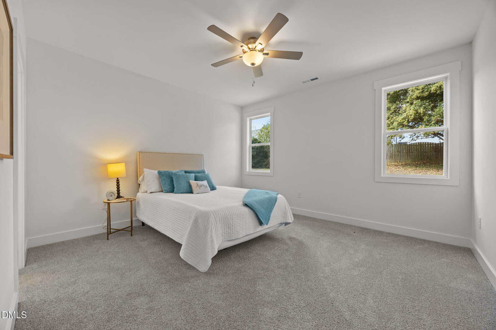 A bright bedroom with gray carpet, white walls, a ceiling fan, and two windows. There's a neatly made bed with white bedding and blue accent pillows, a small bedside table with a lamp, and minimal decor.
