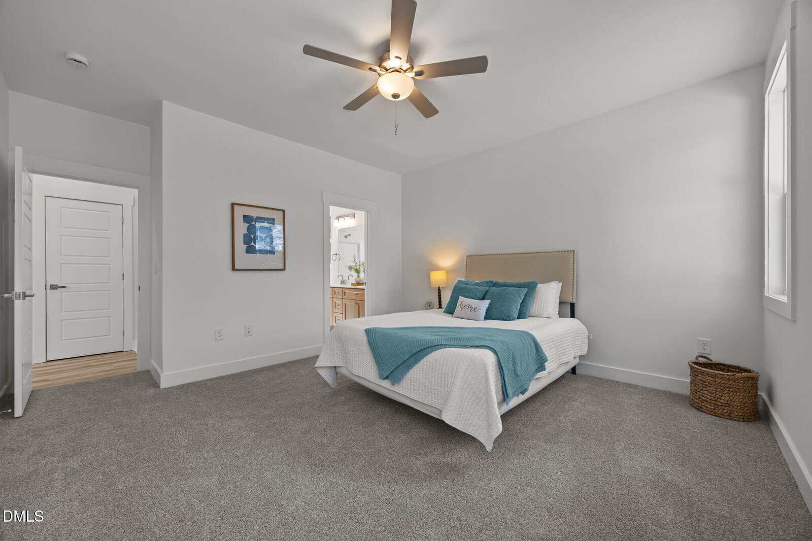 A modern bedroom with gray carpet, a bed with white and teal bedding, a ceiling fan with a light, a nightstand with a lamp, a wall art, and an open door leading to a bathroom. Natural light fills the room.