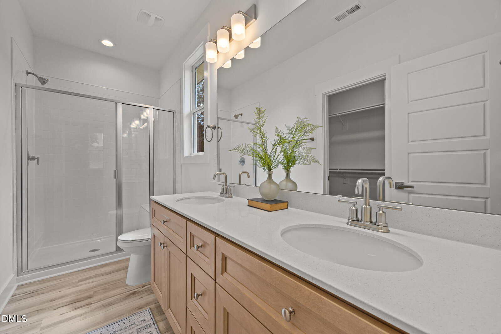 Modern bathroom with a double sink vanity, light wood cabinets, two mirrors, a glass shower, and a white toilet. Greenery in a vase decorates the counter. Light fixtures are mounted above the mirror.