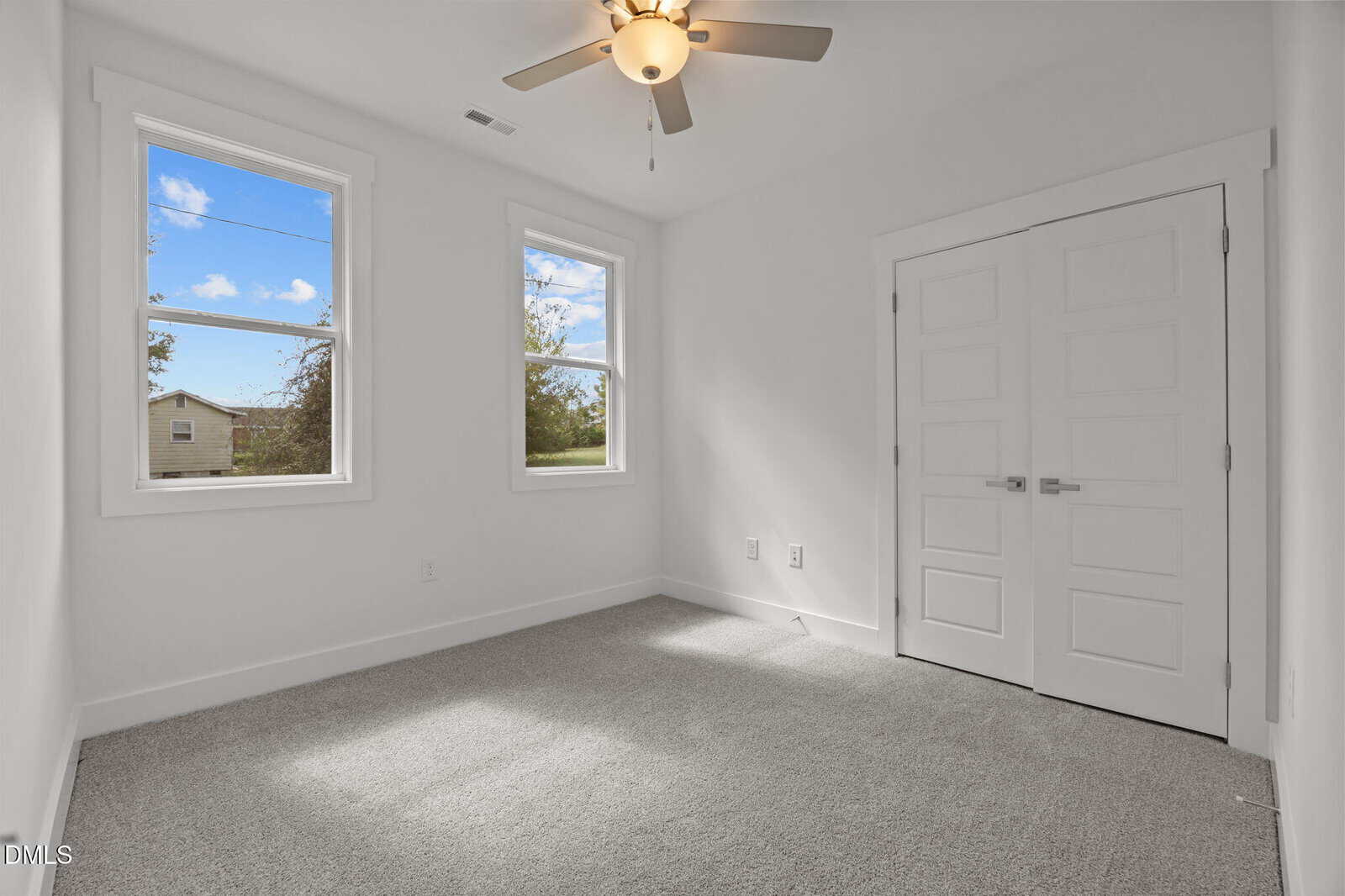 Bright, empty room with white walls, two windows, a ceiling fan, double doors, and light carpeted flooring. Sunlight comes in through the windows, showing a view of trees and a building outside.