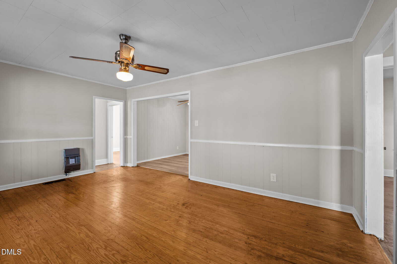 A spacious, empty room with light beige walls, white trim, a wooden floor, ceiling fan with light, and multiple doorways leading to adjacent rooms.