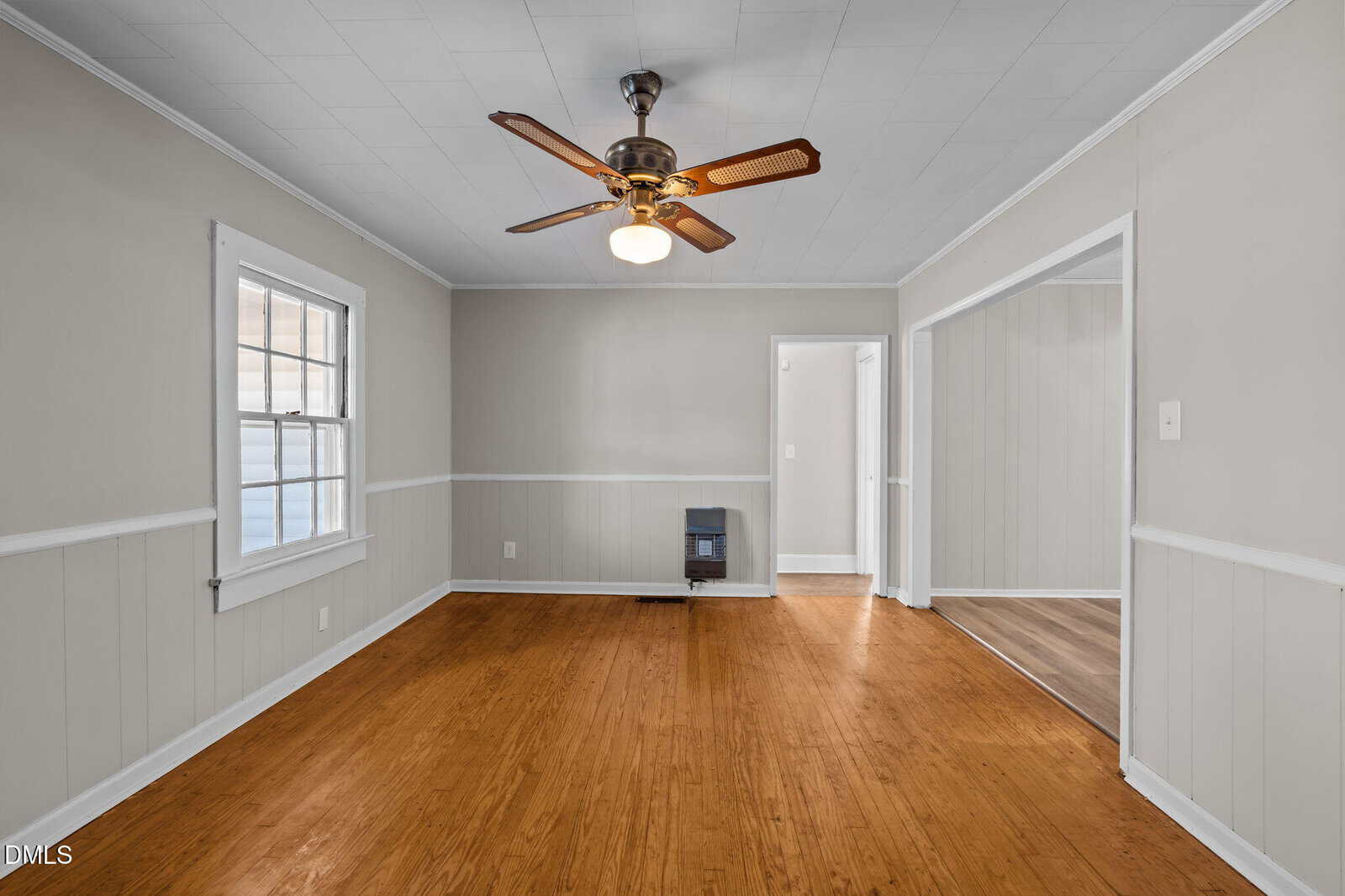 A bright, empty room with wood floors, light gray walls, white trim, a ceiling fan with lights, a window on the left wall, and a small wall heater below. Two doorways lead to other rooms with similar decor.