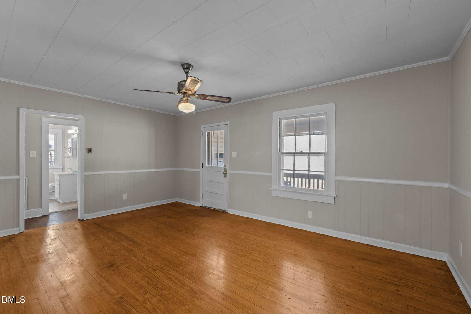 Empty room with wooden floors, beige walls, white trim, a ceiling fan with a light, two windows, and a door leading outside. An open doorway leads to another room in the background.