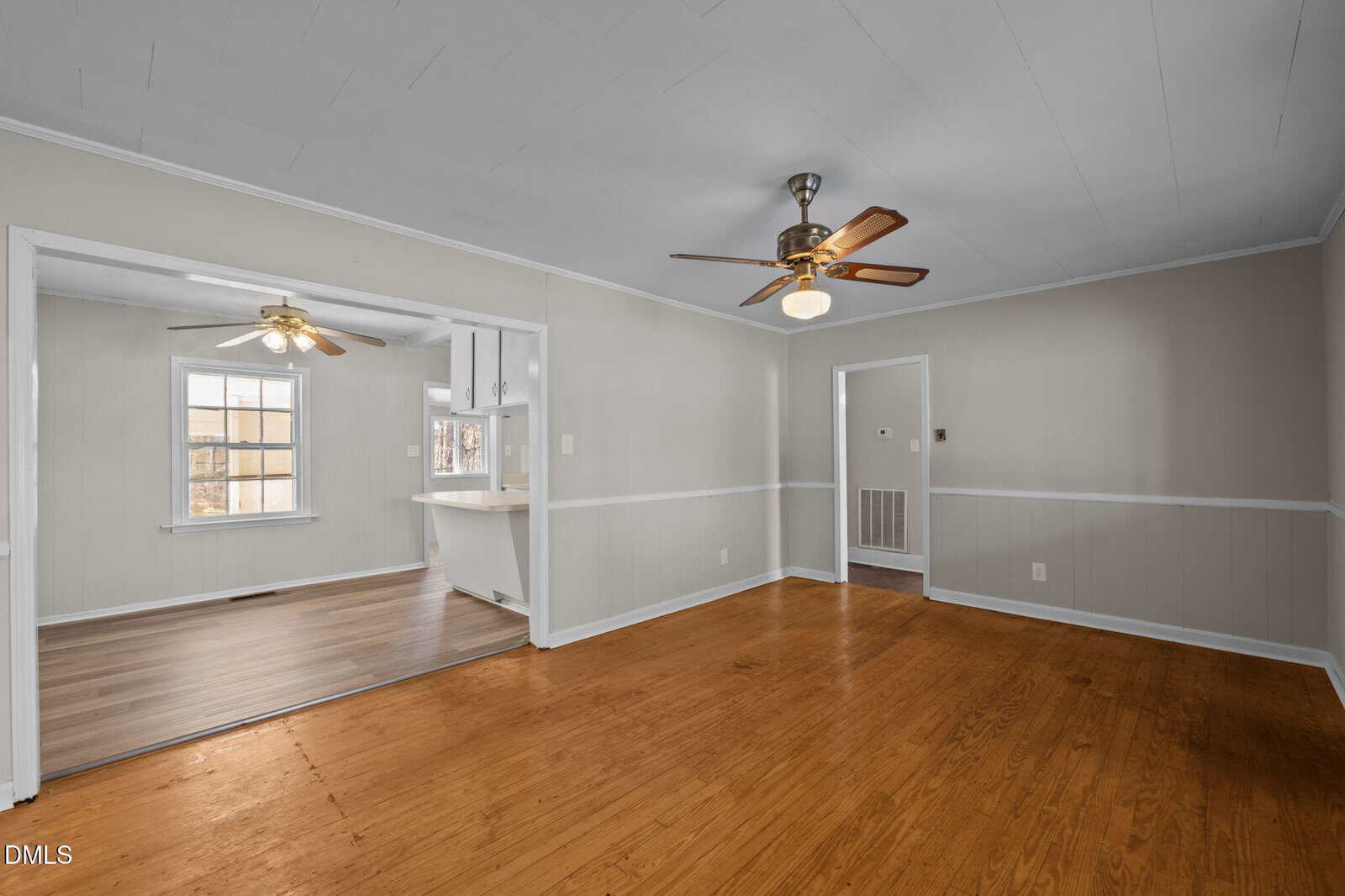 A spacious room with light-colored walls, wood flooring, and a ceiling fan. It opens into a kitchen area with another ceiling fan, white cabinets, and a window letting in natural light.
