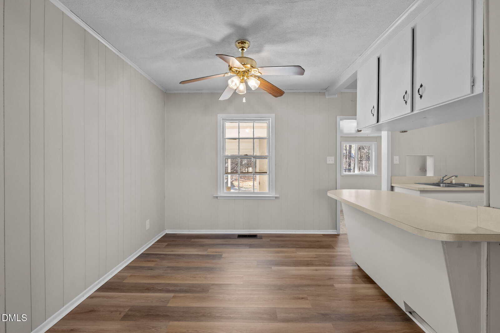 A small, bright kitchen and dining area with light wood flooring, beige paneled walls, a ceiling fan with lights, a window, and white cabinets with a breakfast bar counter.