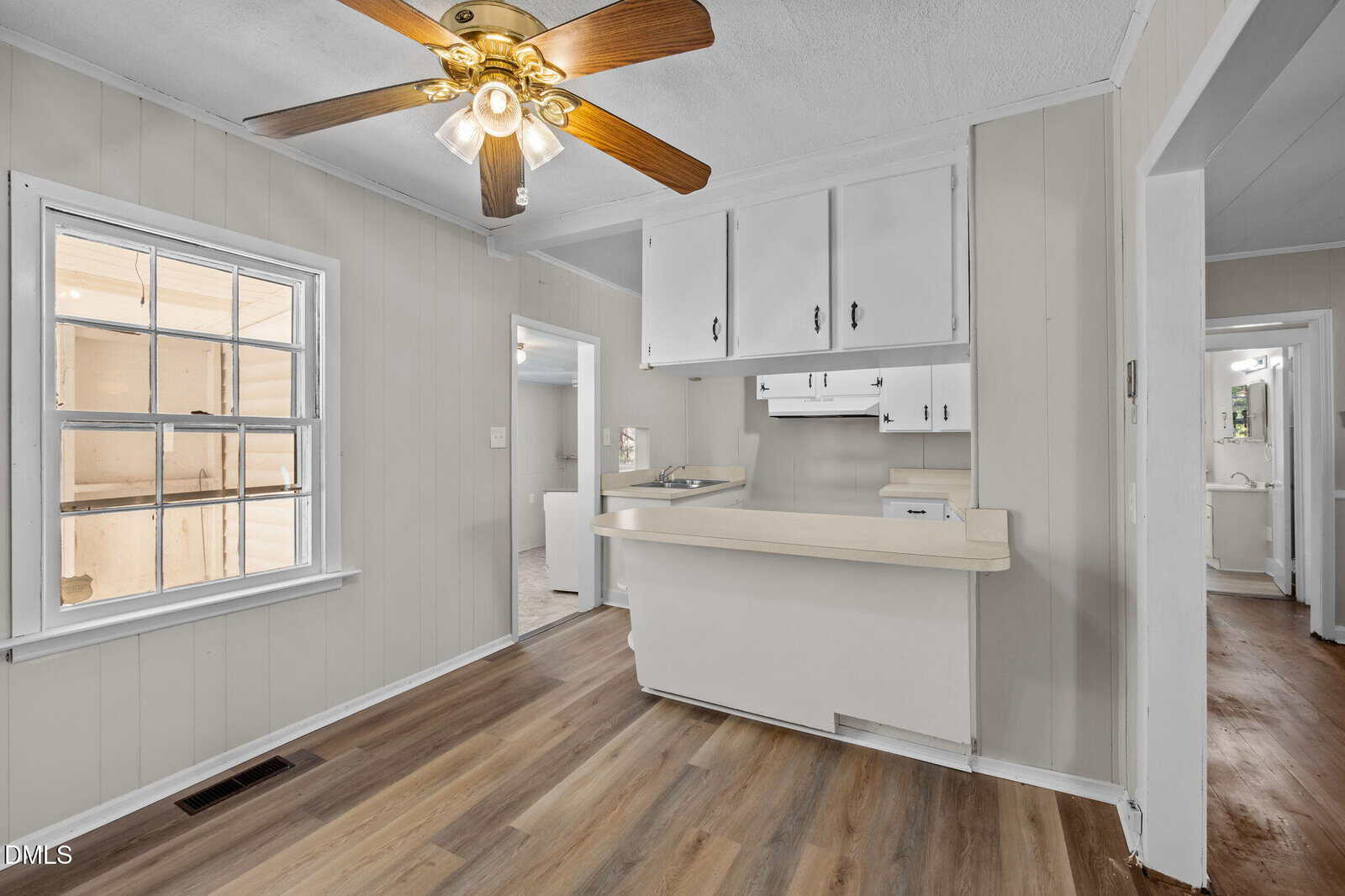 A small kitchen with white cabinets, beige countertops, wood-style flooring, beige paneled walls, and a ceiling fan with lights. A window lets in natural light, and two doorways lead to other rooms.