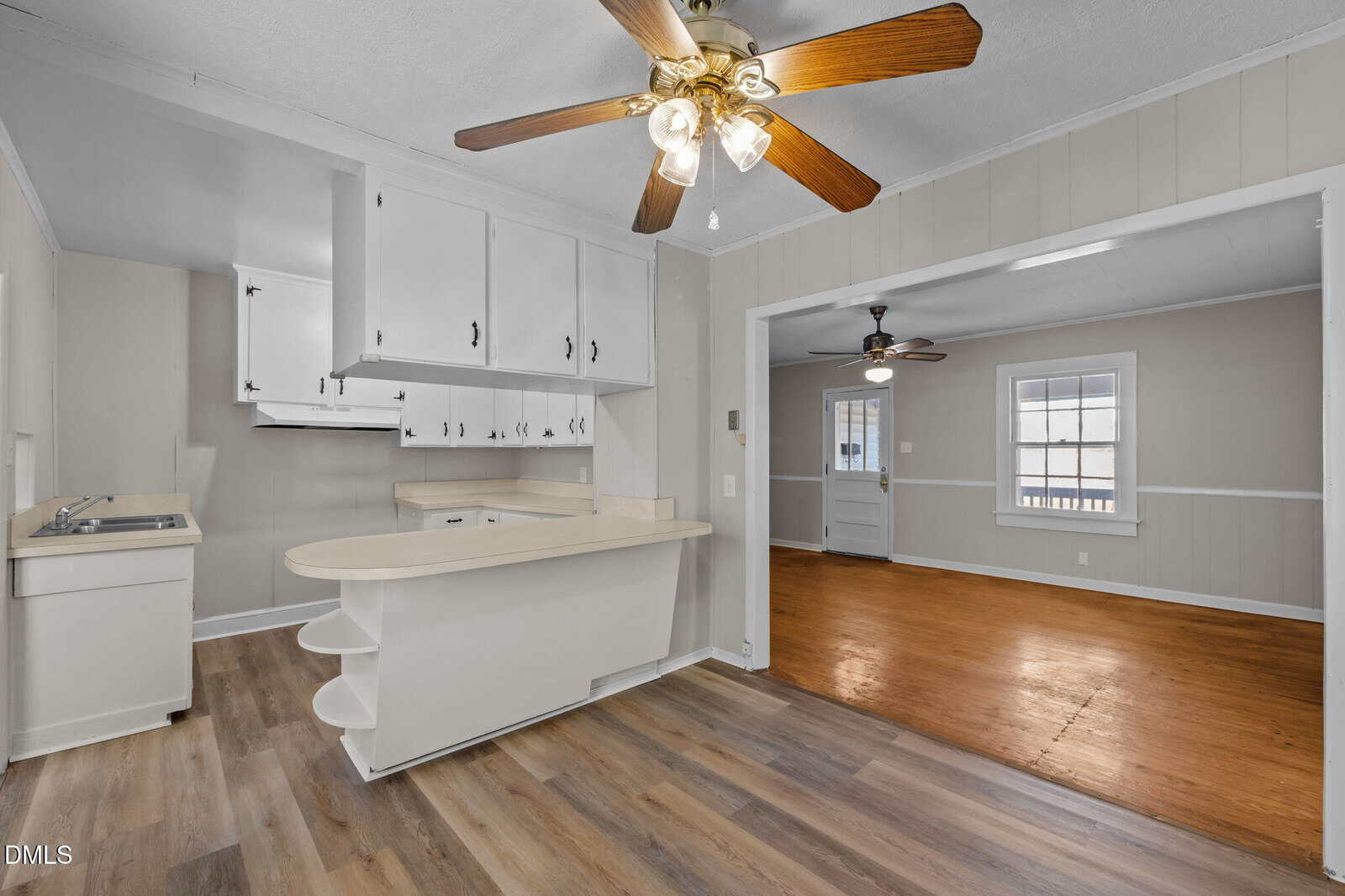 A kitchen with white cabinets, beige countertops, open shelving, and wood-look vinyl flooring. Adjacent is a room with hardwood floors, a ceiling fan, and windows letting in natural light.