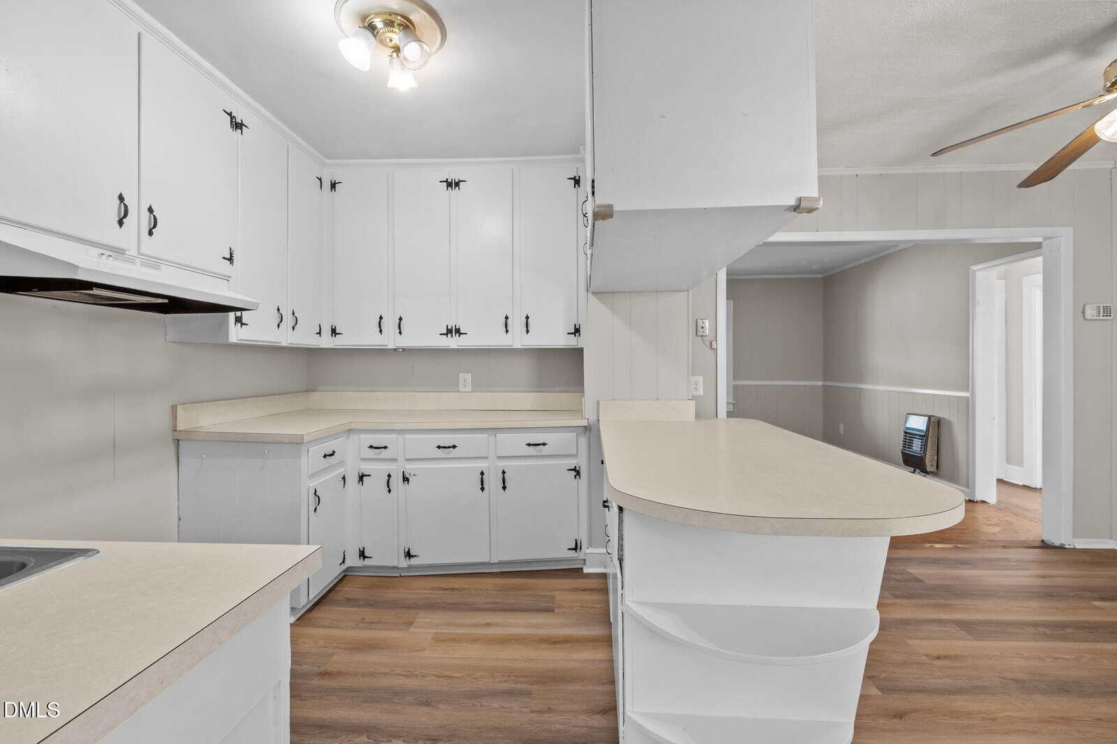 A kitchen with white cabinets, light beige countertops, wood-look flooring, and a ceiling fan. The layout includes upper and lower cabinets, a peninsula counter with open shelving, and an adjacent entry to another room.