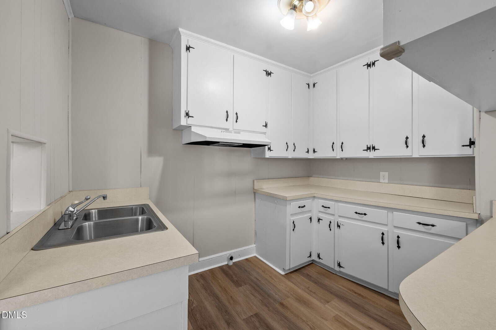 A small kitchen with white cabinets, beige countertops, stainless steel double sink, wood-look flooring, and under-cabinet range hood; walls are a light neutral color.