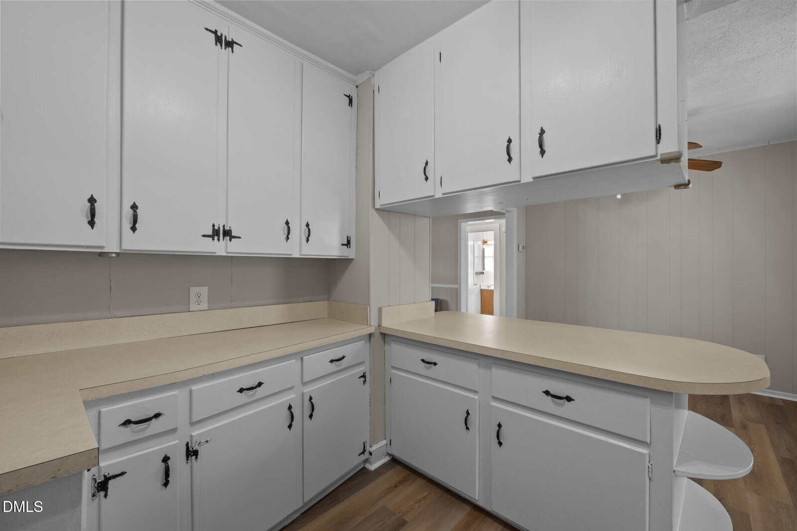 A kitchen with white cabinets, light beige countertops, and wood flooring. The cabinets have black handles, and there is an L-shaped counter with space for seating. Walls are light-colored.