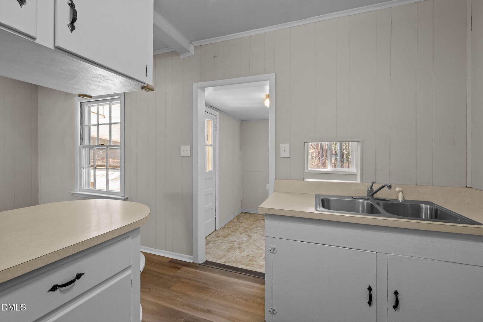Small kitchen with white cabinets, light countertops, double sink, wood flooring, and beige paneled walls. Two windows and a doorway lead to a bright adjacent room with a white door and patterned linoleum floor.
