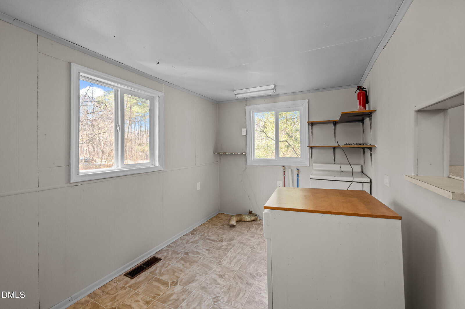 A small, empty room with two windows, white walls, shelves, a wooden counter, tile floor, and a fire extinguisher on the wall. Light streams in from the windows, and trees are visible outside.