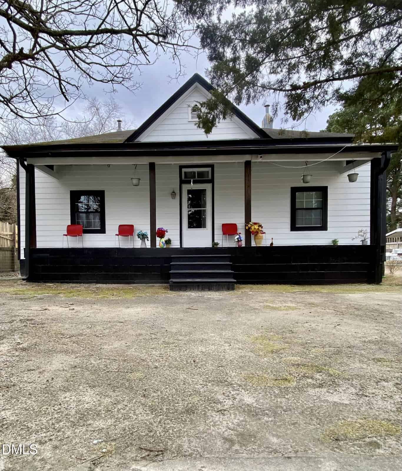A white house with black trim at 332 Chavasse Avenue in Henderson features a covered front porch with two red chairs, potted plants, and three steps leading up from a wide concrete driveway. Trees border the yard under a cloudy sky.