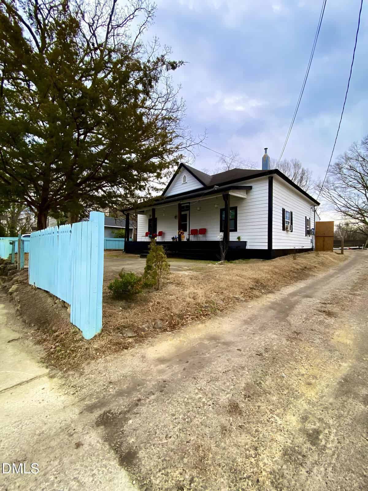 A small white house with black trim and a covered porch featuring red chairs, sits beside a dirt driveway. A turquoise wooden fence lines the front yard, with bare trees and a cloudy sky in the background.