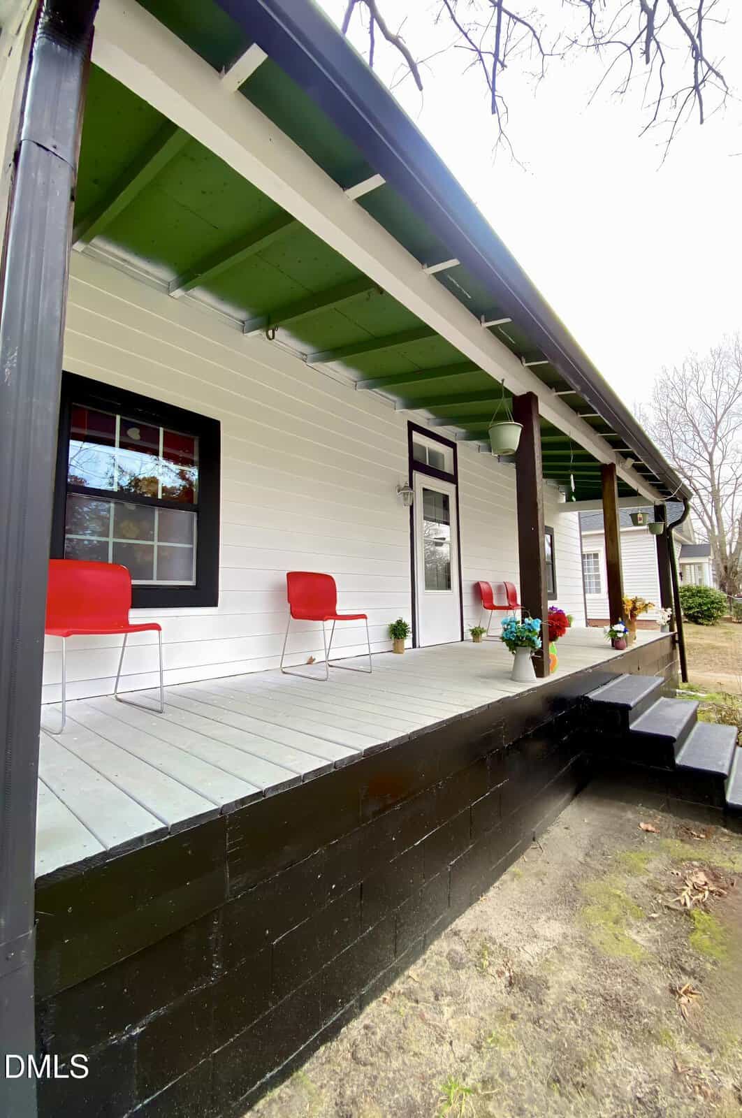 A white house with a green ceilinged front porch, black trim, and two red chairs beside potted plants. The porch has wide steps and is surrounded by bare trees and a grassy yard.