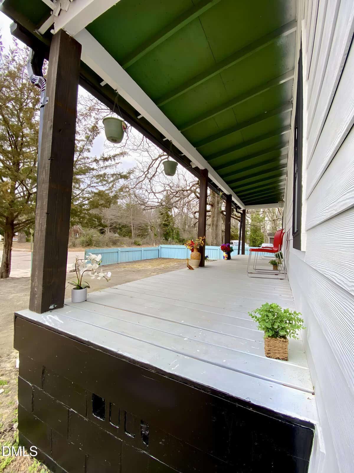 A wide view of a porch with a green ceiling, white floor, black posts, and potted plants. Two red chairs and a small table are near the door. Trees and a blue fence line the yard in the background.