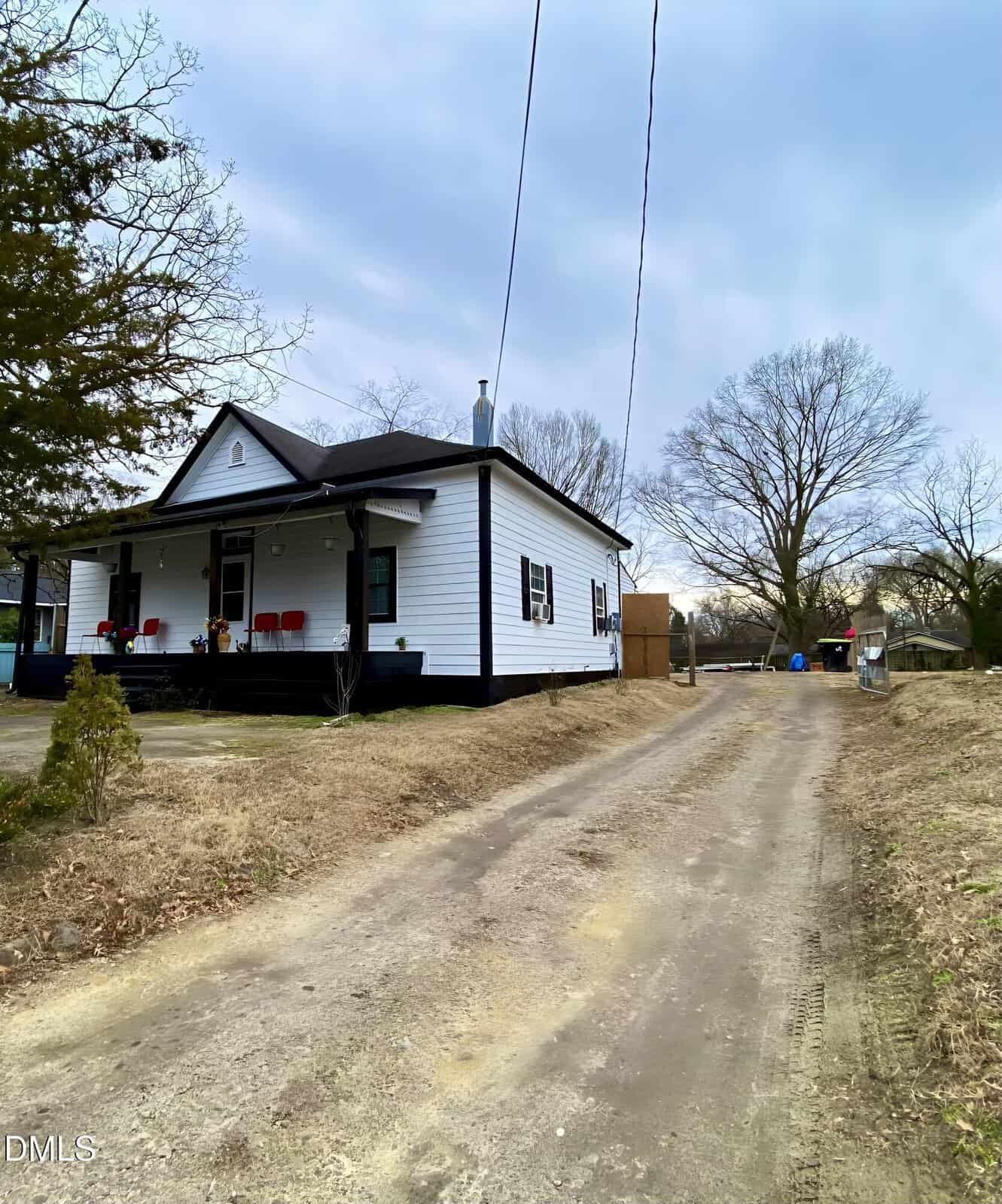 A white, single-story house with a covered porch and red chairs sits beside a long dirt driveway, surrounded by leafless trees and patchy grass under a cloudy sky.