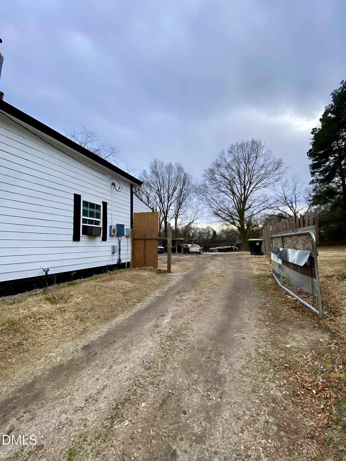 A dirt driveway runs alongside a white house with black shutters on the left. Leafless trees and a wooden fence are on the right. The sky is cloudy and overcast. A metal gate is partially open near the driveway entrance.