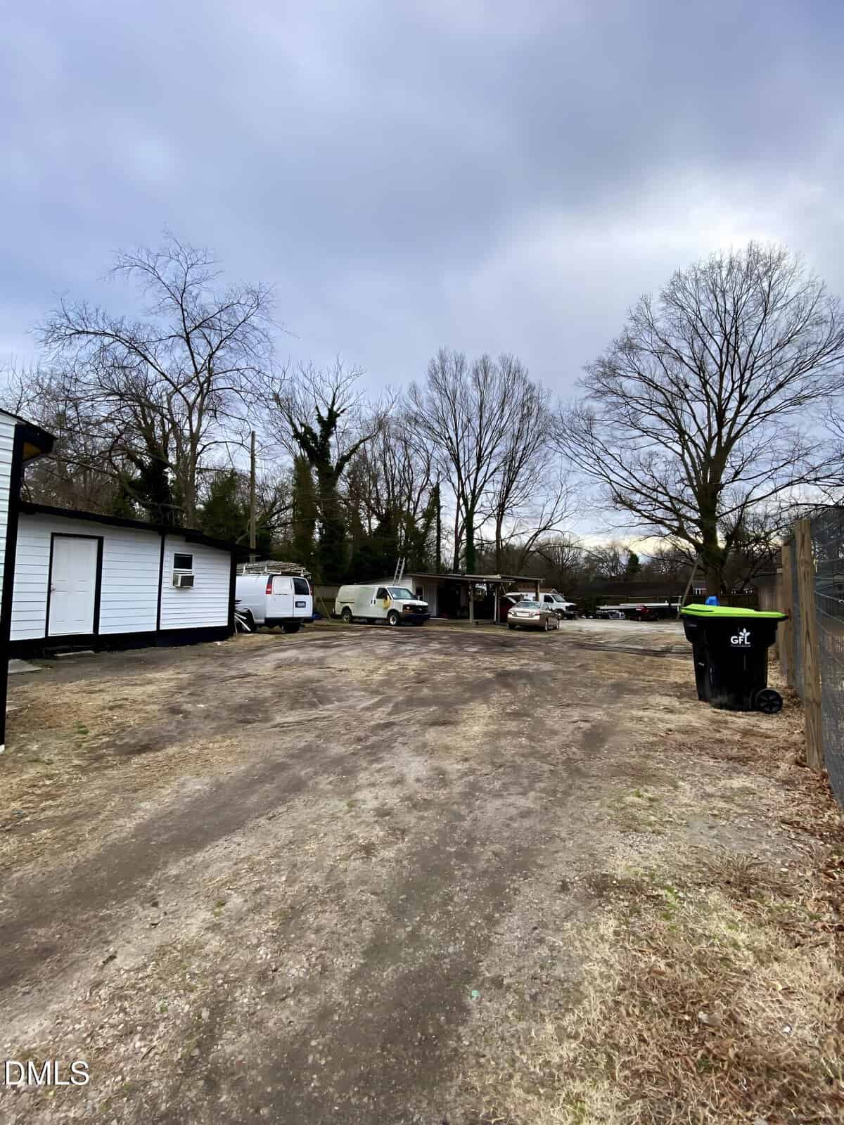 A wide, dirt driveway leads to a small white building on the left, with several vehicles and trees without leaves in the background under an overcast sky. Trash bins are visible near a wooden fence on the right.