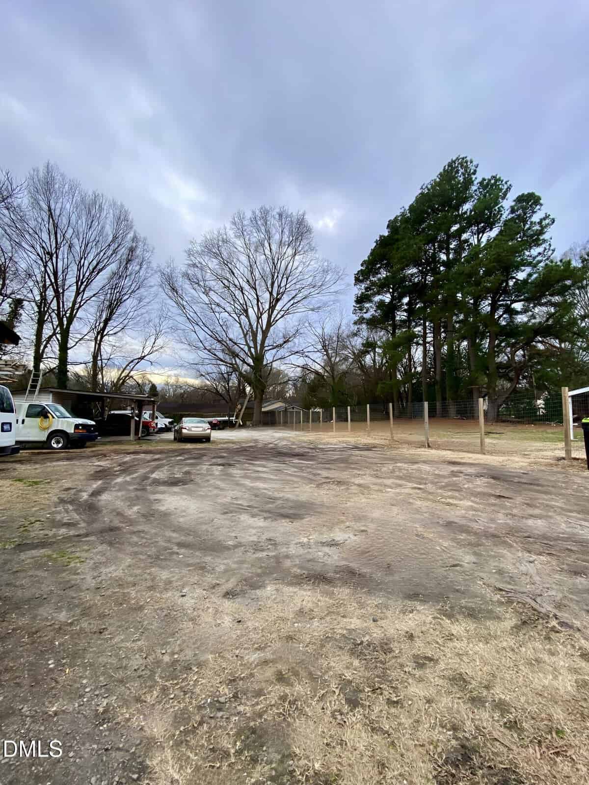 A wide, open dirt and grass lot with a few parked vehicles, surrounded by leafless and evergreen trees, under a cloudy sky. Some houses and a chain-link fence are visible in the background.
