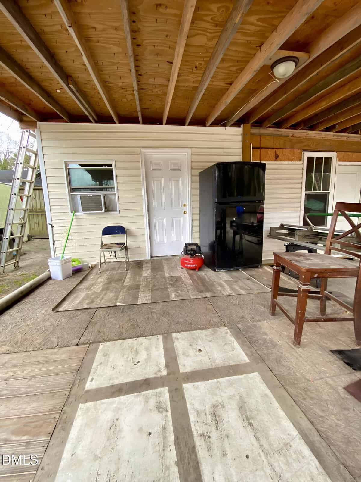Covered porch with exposed wooden beams, a white door, a black refrigerator, a folding chair, cleaning supplies, a red tool case, a wooden bench, and various items scattered on a wood plank floor.