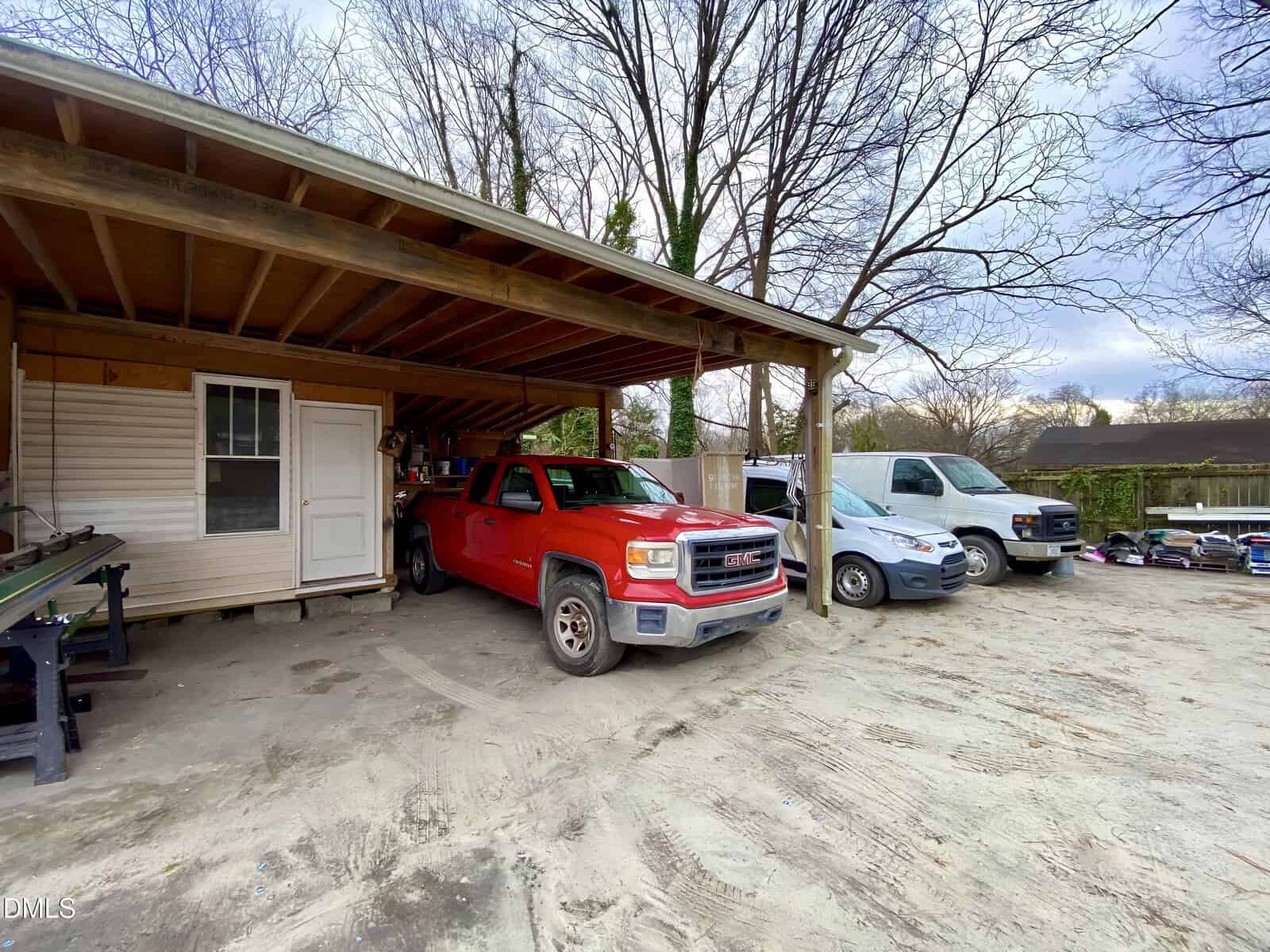 A red pickup truck and two vans are parked under a wooden carport attached to a small building, with leafless trees and other vehicles in the background.