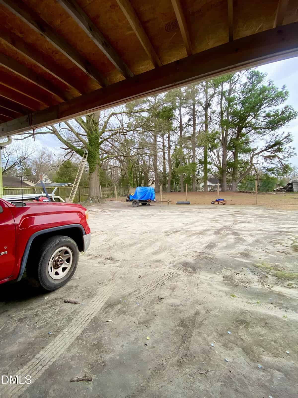 View of a sandy, open yard with a red truck on the left, a blue tarp-covered trailer in the center, scattered items, a ladder, and tall trees in the background, partially shaded by a wooden roof.