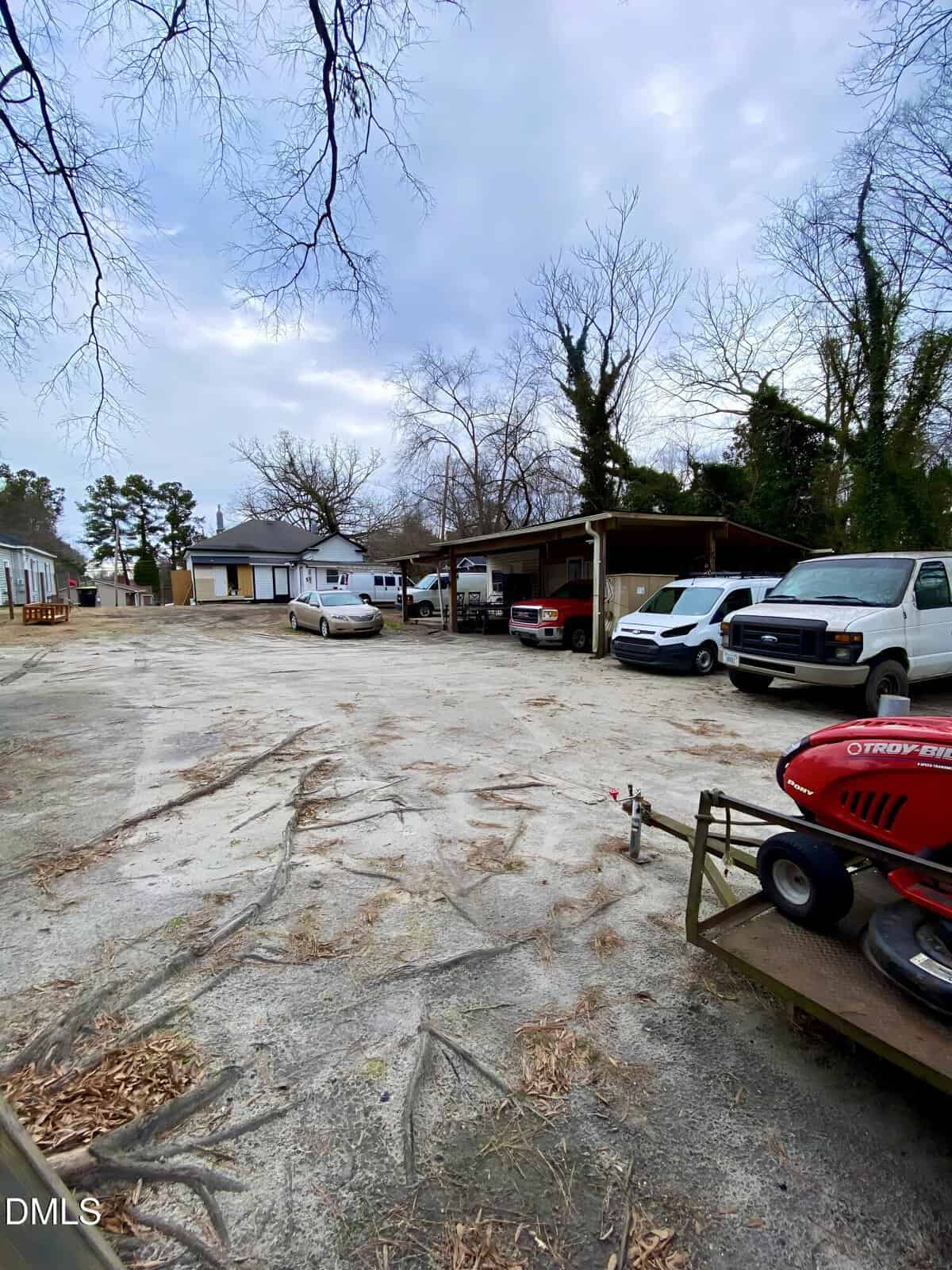 A sandy, open lot with several parked vehicles, carports, and a white house in the background. Bare trees surround the area, and a red riding lawnmower is in the foreground on the right.