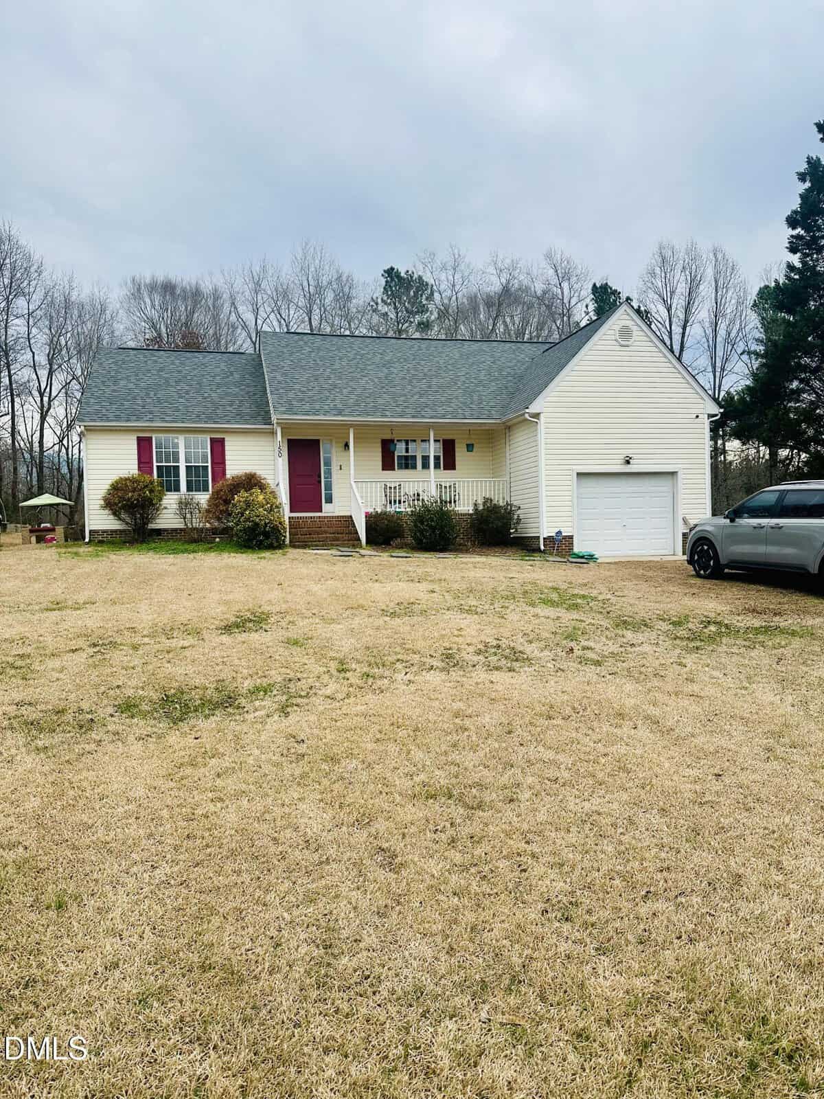 A single-story light yellow house with a front porch, red shutters, and a garage sits on a large, grassy lawn. A gray SUV is parked on the driveway, and bare trees are in the background under an overcast sky.