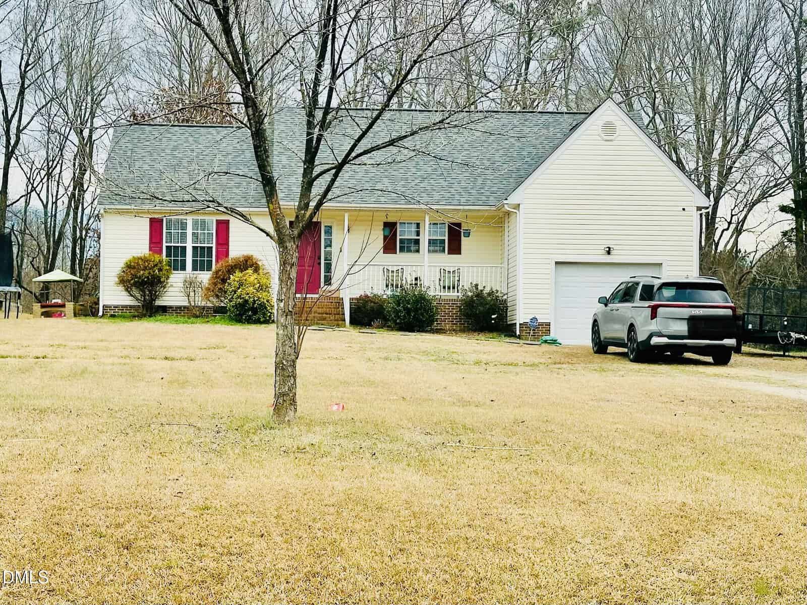 A light yellow single-story house with red shutters, a front porch, and attached garage sits on a large grassy yard. A gray SUV is parked in the driveway, and leafless trees are in the foreground.