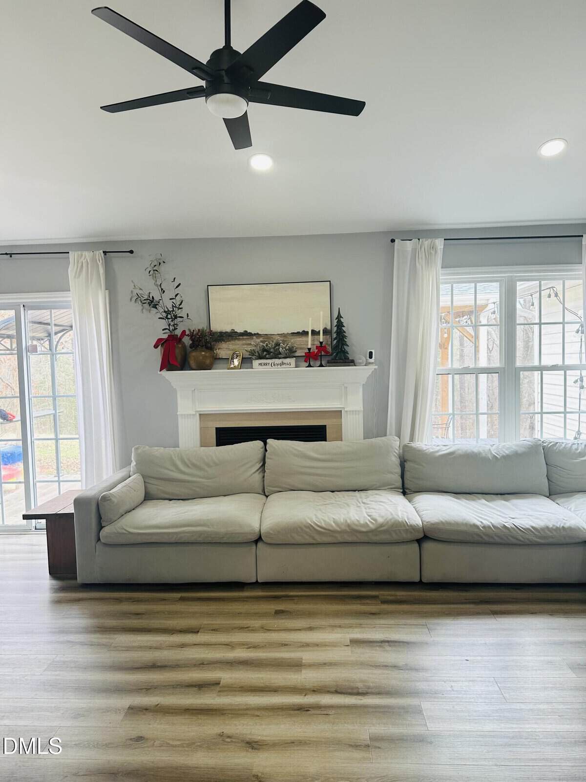 A bright living room with a large beige sofa in front of a white fireplace. The mantel is decorated with greenery, red bows, candles, and a framed landscape. Tall windows with white curtains let in natural light.