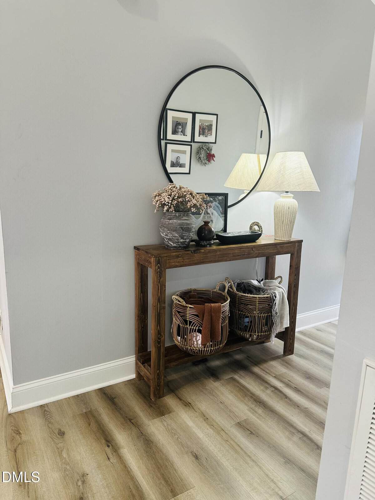 A wooden console table with two wicker baskets sits against a light gray wall. Above it hangs a round mirror reflecting framed photos, with a lamp, vase of dried flowers, and decorative items on the table.