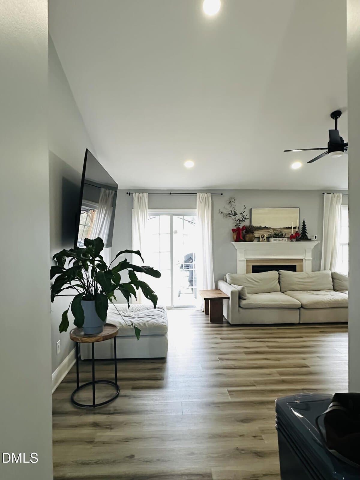 Bright living room with light wood floors, a white sectional sofa, a fireplace with decor, a potted plant on a small table, and large windows with white curtains letting in natural light.
