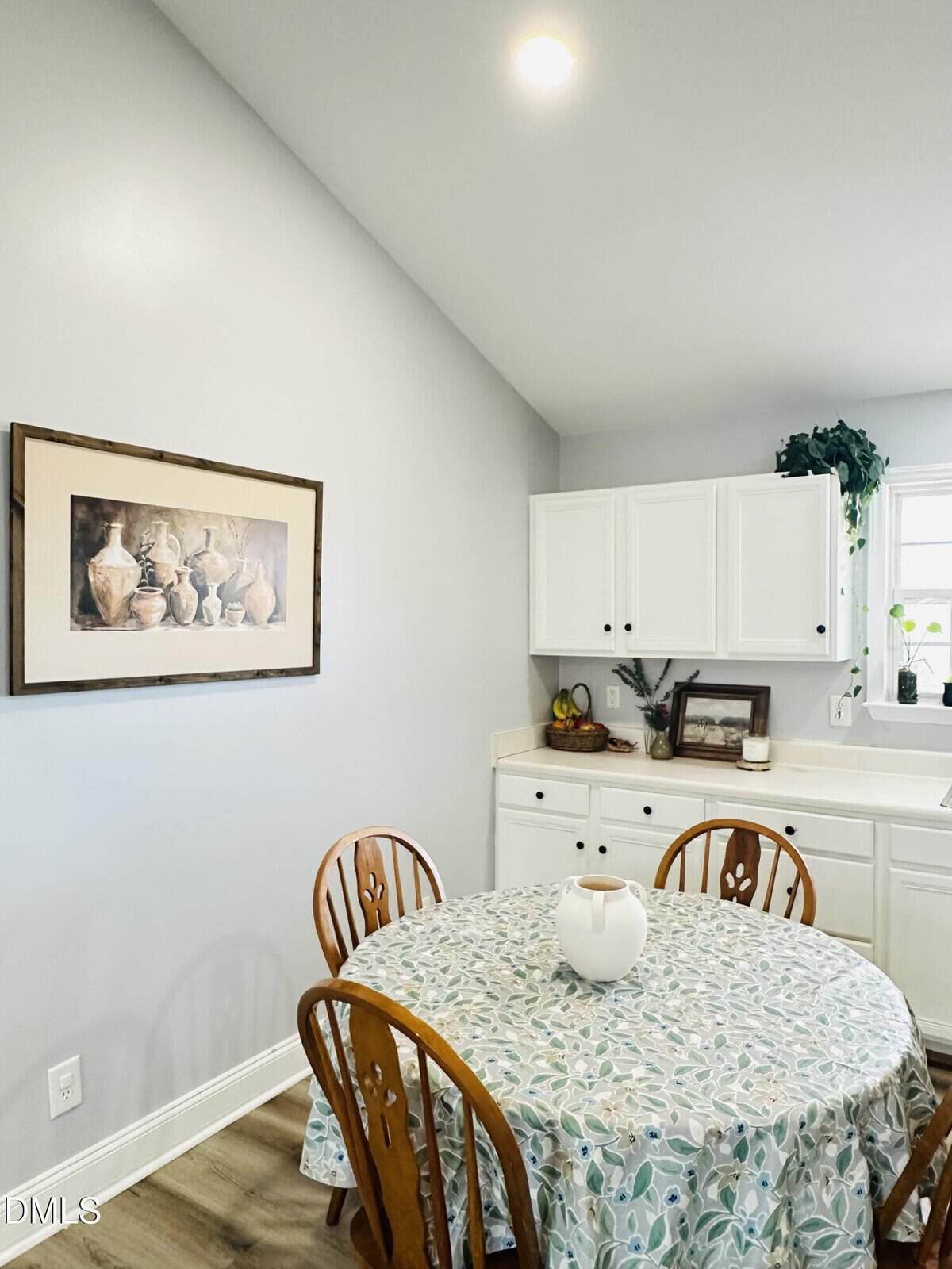 A small, bright kitchen with a round table covered by a patterned tablecloth, wooden chairs, white cabinets, a window with plants, and a framed still life painting on a light gray wall.