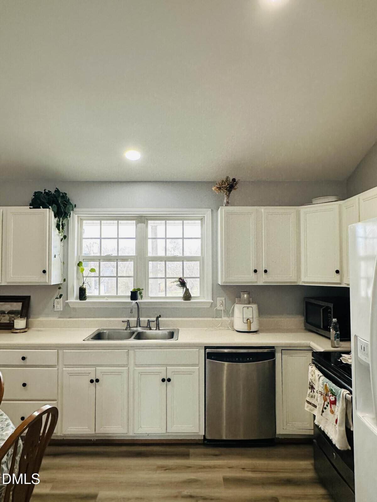 A bright kitchen with white cabinets, a double sink under two windows, a dishwasher, stove, microwave, and refrigerator. Several plants and decor items sit on the windowsill and counters.