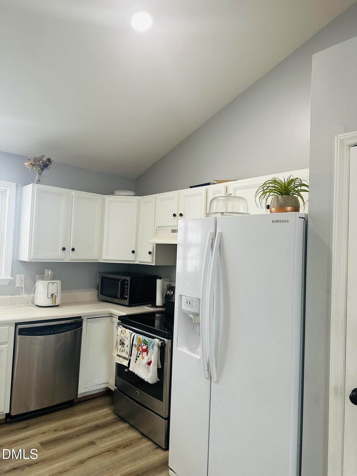 A modern kitchen with white cabinets, stainless steel appliances, a white refrigerator, light wood flooring, and small plants on top of the fridge and cabinets. Bright natural light comes from a window on the left.