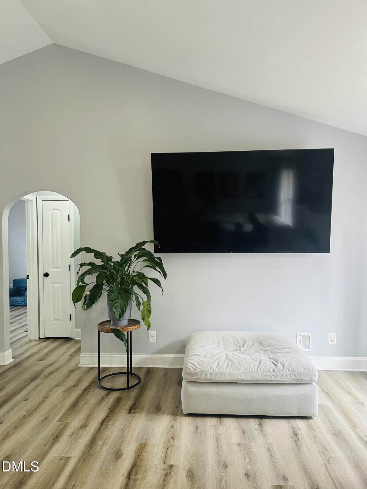 A modern living space with light wood flooring, a large wall-mounted flat-screen TV, a white ottoman, a potted plant on a small round table, and an arched doorway leading to another room.