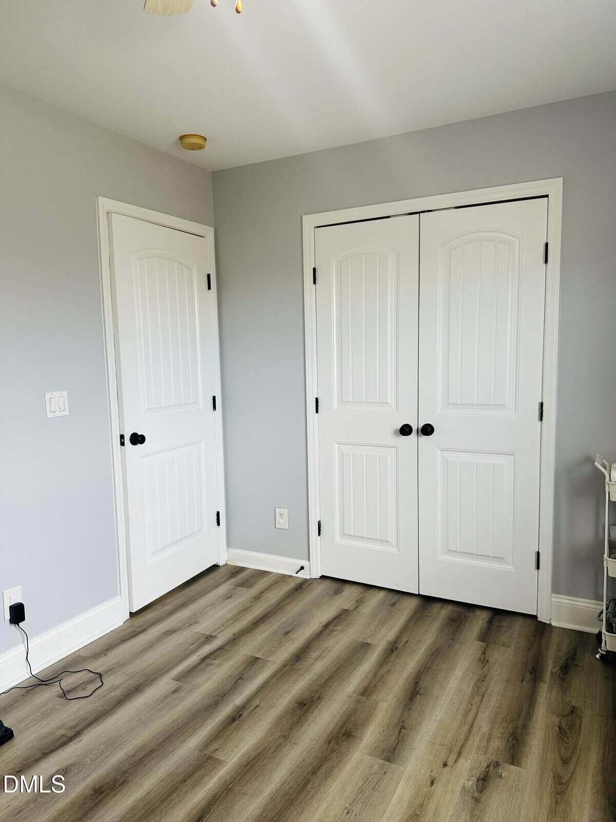A bright, empty room with light gray walls, wood flooring, and two white doors—one single and one double. There are no furnishings except a small white shelf in the corner.