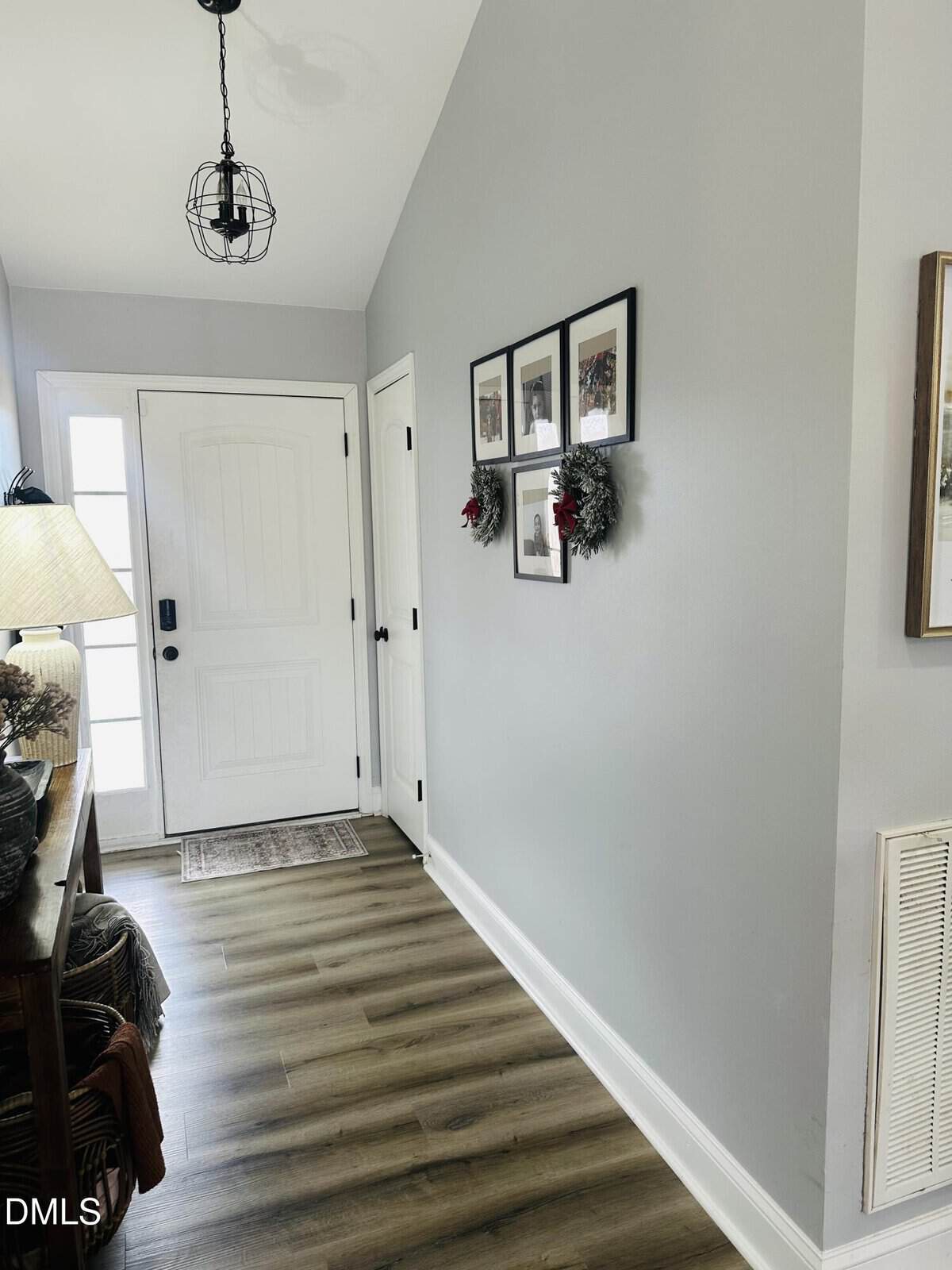 A hallway with light gray walls, wood flooring, a white front door, framed photos, and small wreaths on the wall. A console table with a lamp and decor sits on the left. Natural light comes through the door.