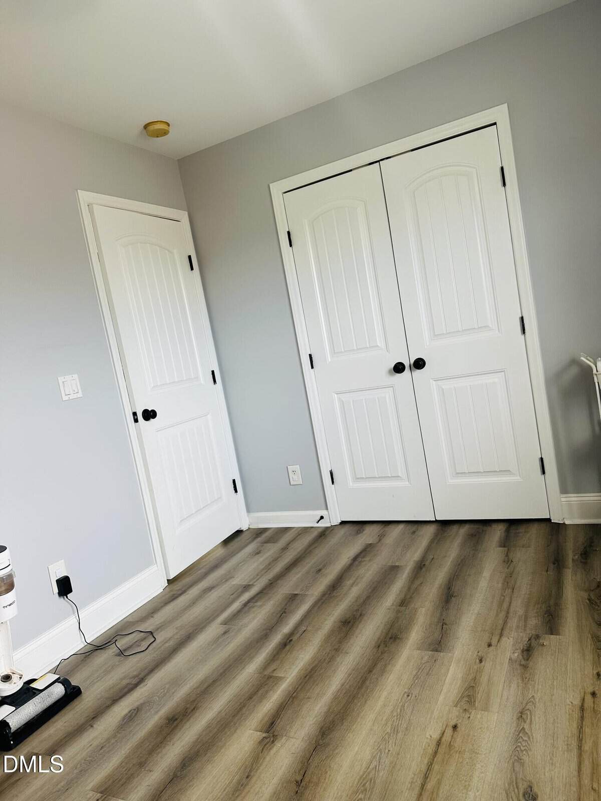 A bright room with light gray walls, a wooden floor, two white doors, one closet door, and a vacuum cleaner in the corner near an electrical outlet.
